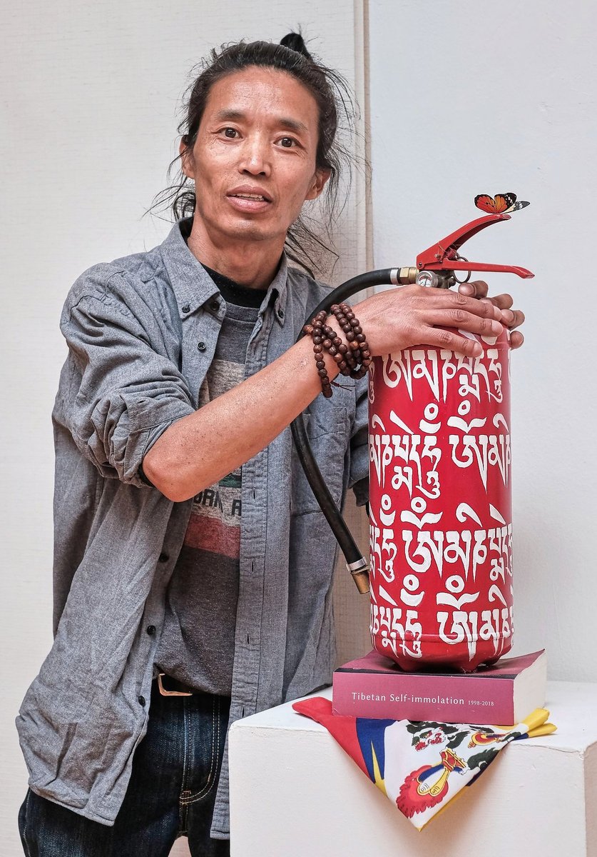 Chungpo Tsering s hasicím přístrojem nesoucím mantru „O mani padme hum", tibetskou vlajkou a knihou Tibetan Self-immolation 1998–2018